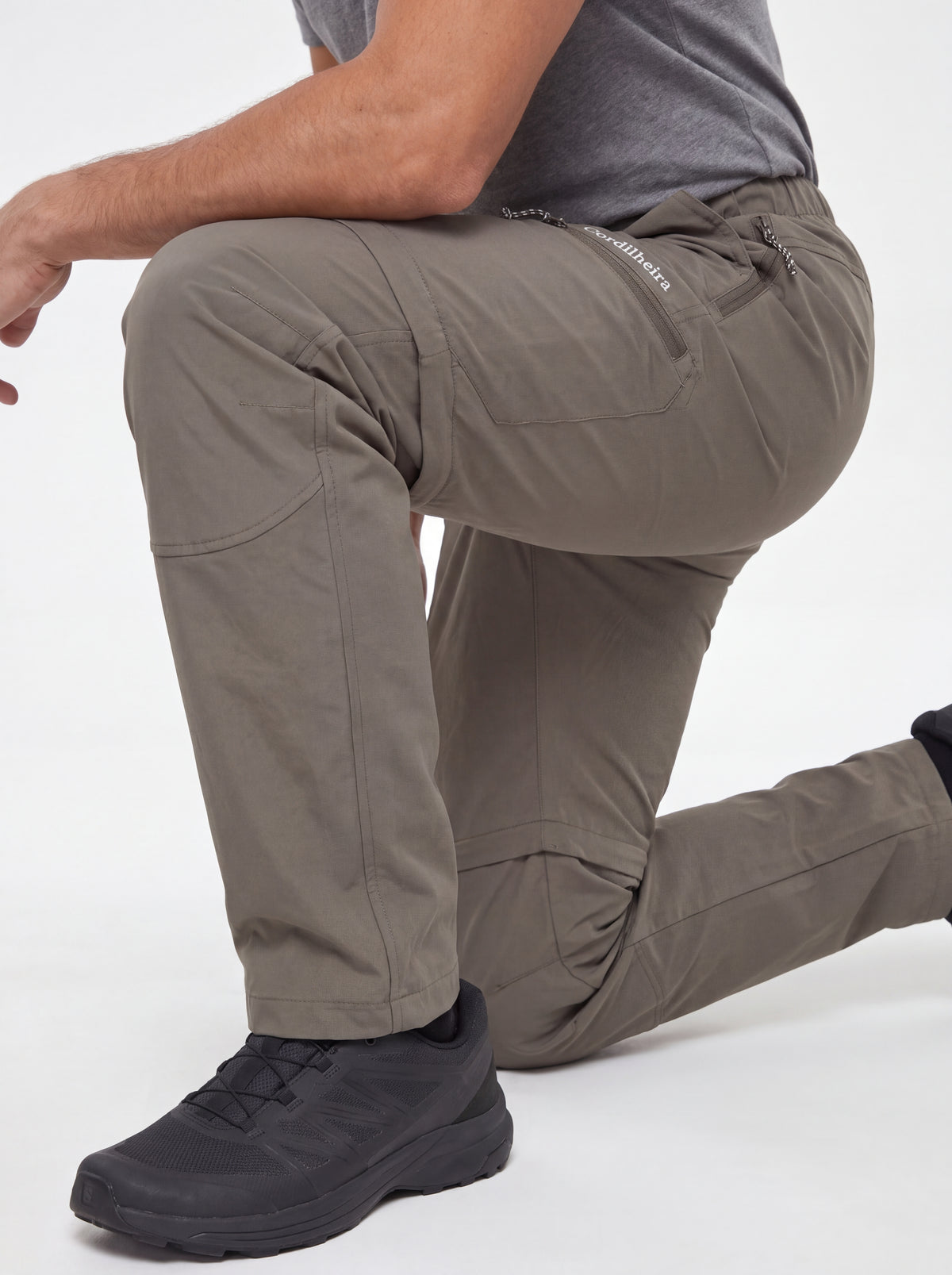 Modelo ajoelhado usando Calça 2 em 1 Cordilheira Canopy Masculina com logo em destaque, Roupa da Marca Cordilheira com Boa mobilidade, resistência e qualidade Confiável para trilhas.